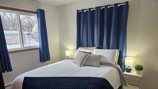 Un dormitorio ordenado con cama, lámparas de noche, una planta pequeña y vista al exterior.