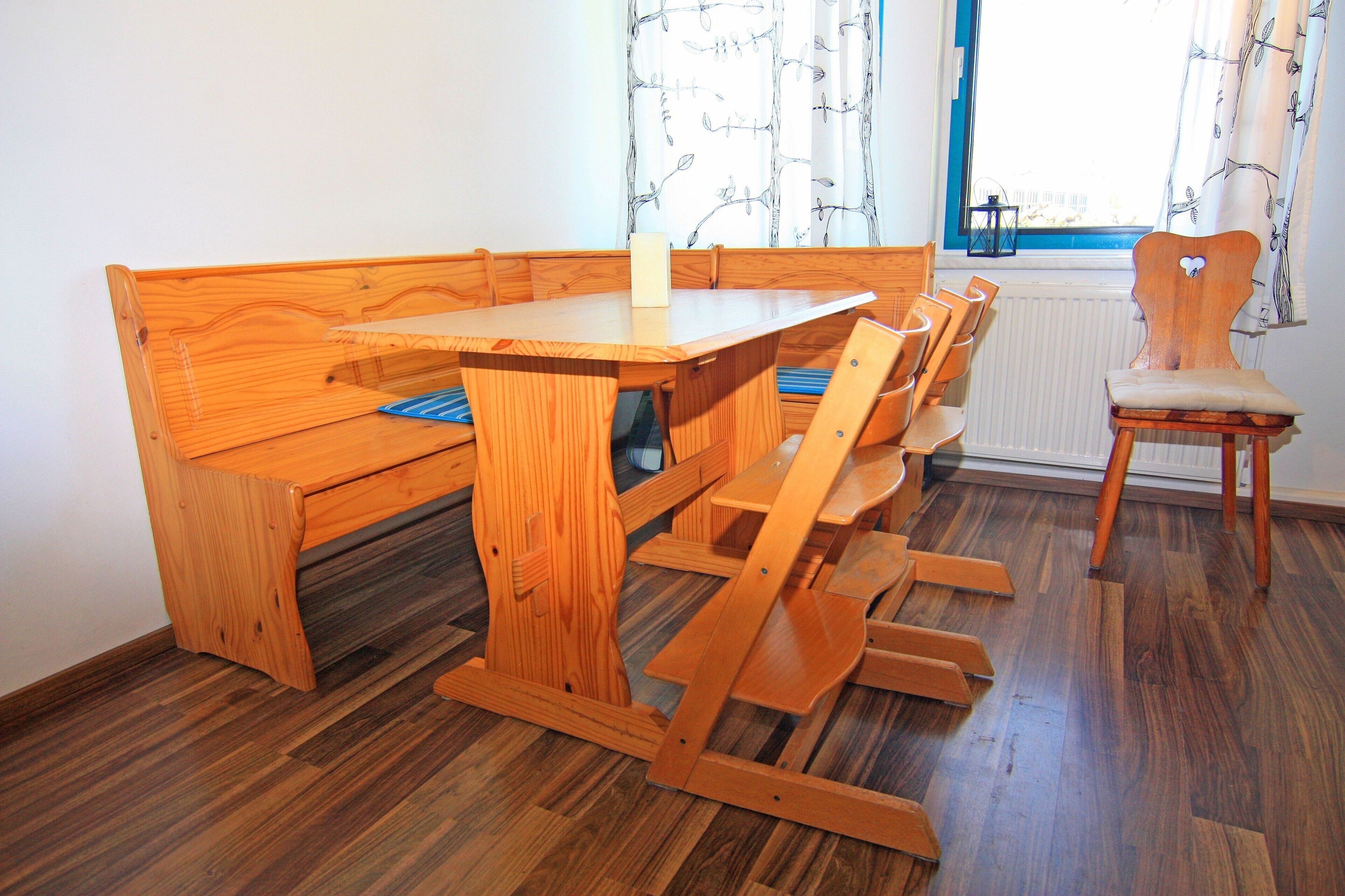 A wooden dining table with benches, a chair, and a radiator in a room with wooden flooring and a window with curtains.