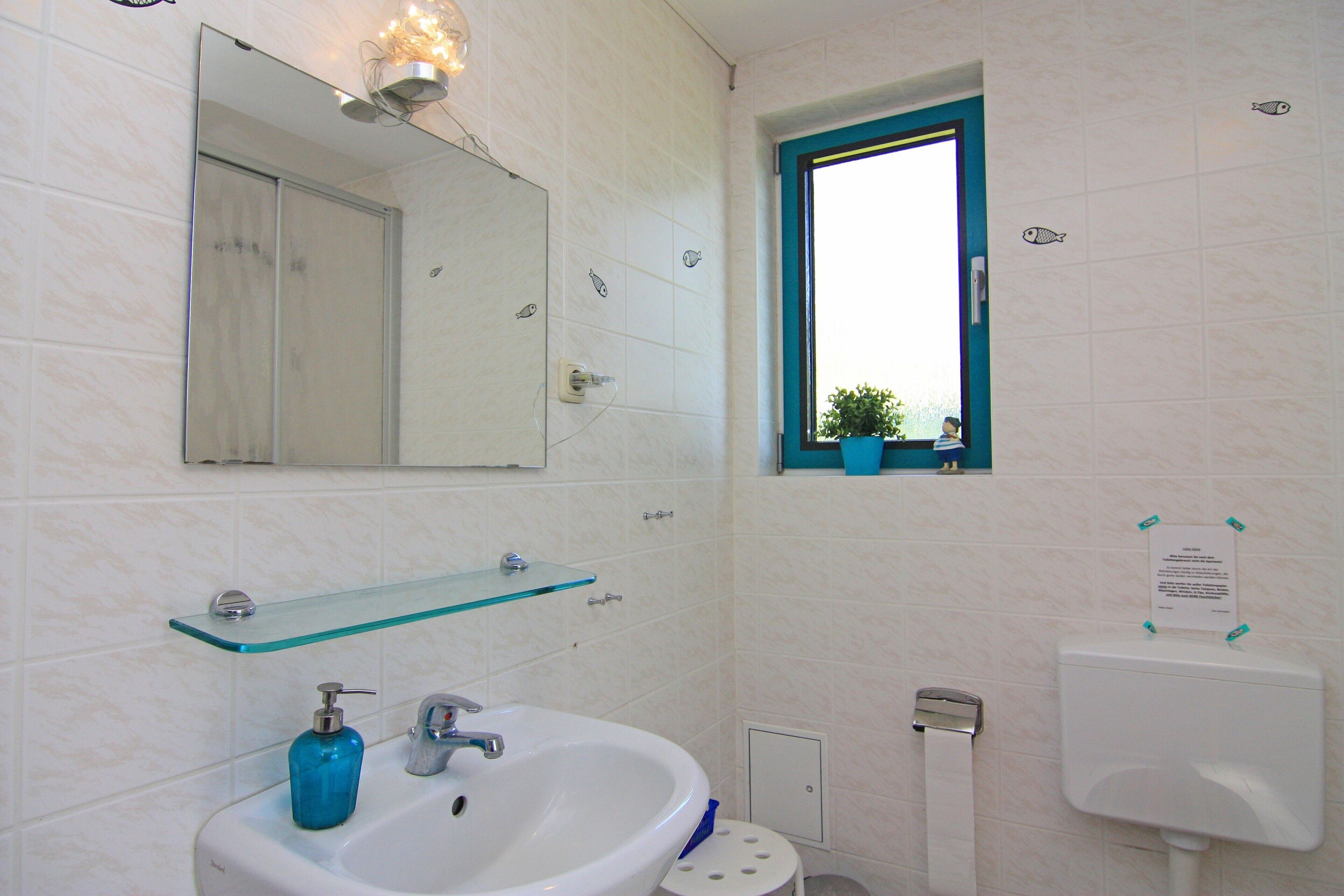 A bathroom with a white sink, a glass shelf, a mirror, a window, and a toilet.