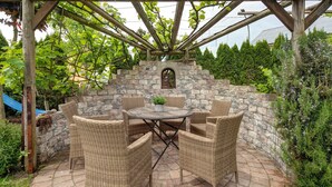 A covered outdoor seating area with wicker chairs and a table, surrounded by stone walls and greenery.