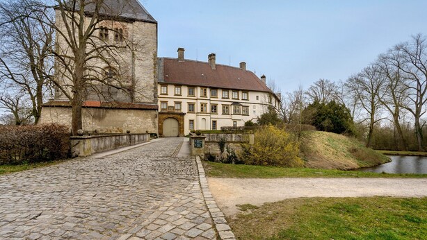 Un bâtiment historique, avec un chemin pavé menant à une entrée fermée par un portail, entouré d’arbres et bordé d’une  étendue d’eau.