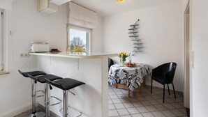 Une cuisine avec une fenêtre, une table recouverte d’une nappe à motifs et deux chaises noires.