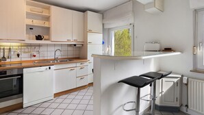 Une cuisine moderne avec des meubles blancs, un lave-vaisselle, un four encastré, un plan de travail en verre et un tabouret de bar.