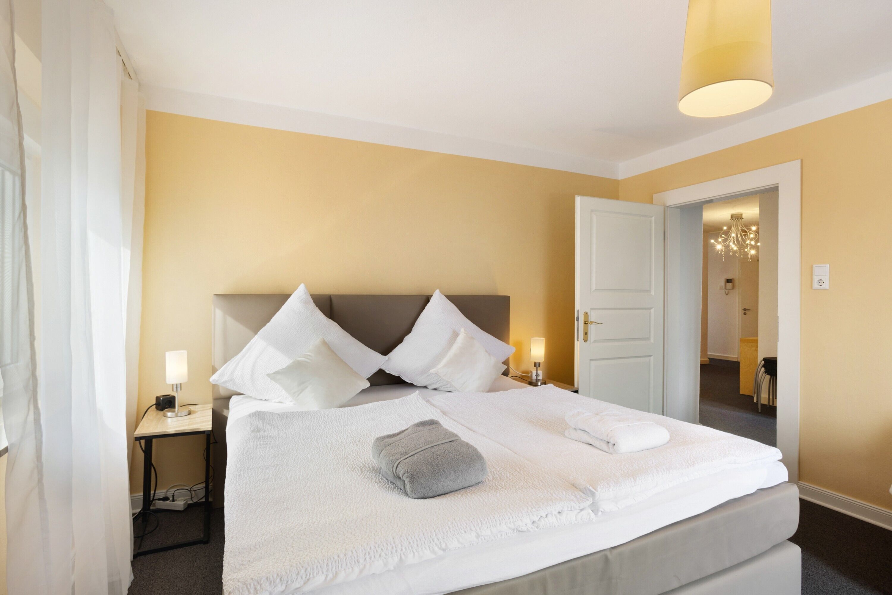 Ein Hotelzimmer mit Bett, weißen Laken, einem Nachttisch mit Lampe und einer Tür zu einem weiteren Raum.