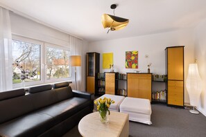 Un salon moderne avec un canapé noir, un pouf blanc, une table basse ornée d’un vase de fleurs, une bibliothèque, un meuble en bois et une grande fenêtre.