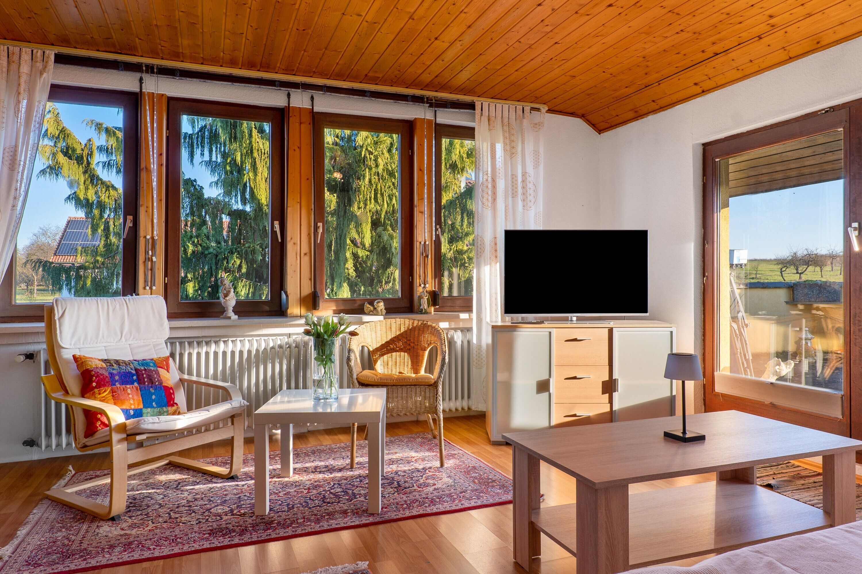 Une pièce avec un plafond en bois, de grandes fenêtres avec des rideaux, une télévision, un fauteuil en osier, une table et un radiateur.