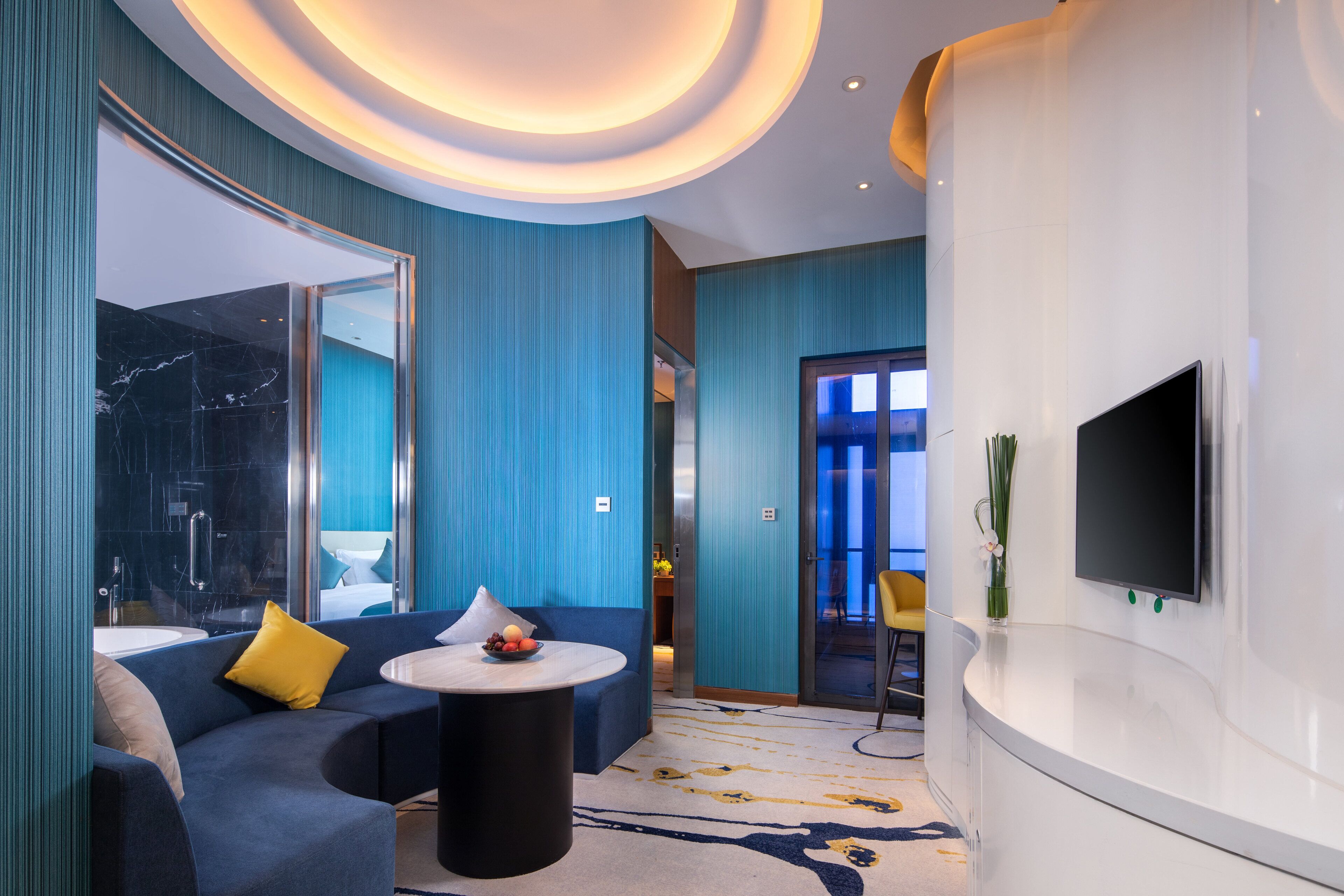 Quarto de hotel moderno com decoração azul, uma mesa redonda com frutas, um sofá com almofadas amarelas, uma TV fixada na parede e um espelho grande.