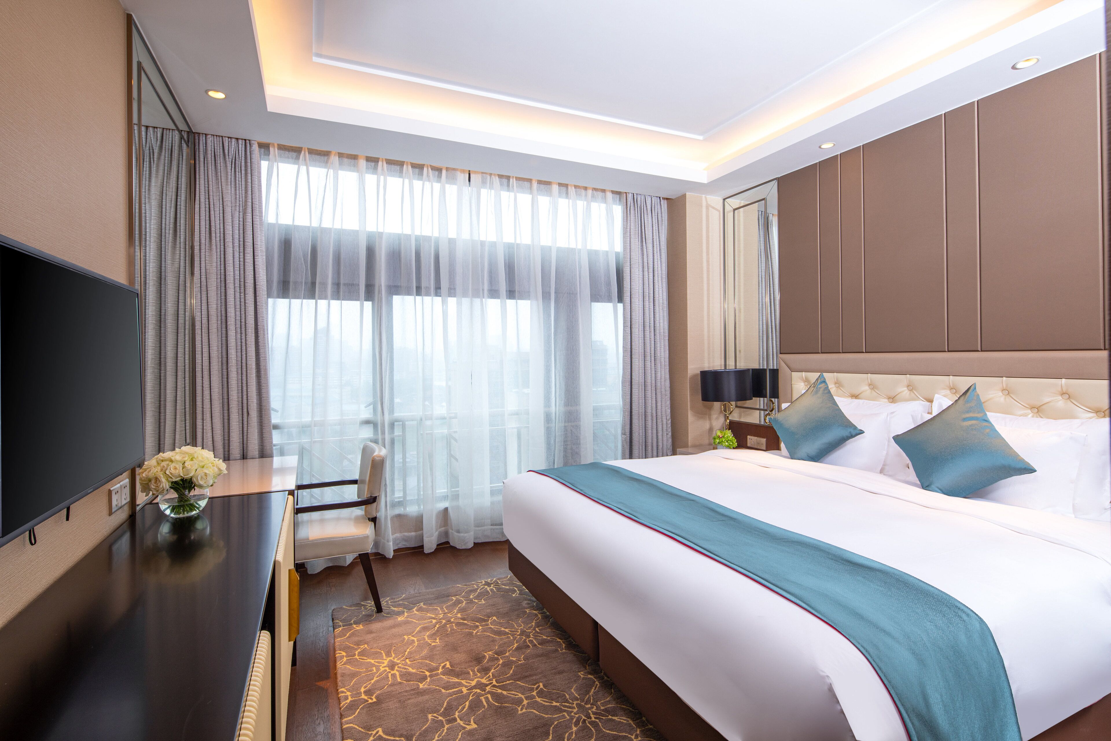 A modern hotel room with a large bed, a flat-screen TV, a desk with a vase of flowers, and a view of the city through large windows.