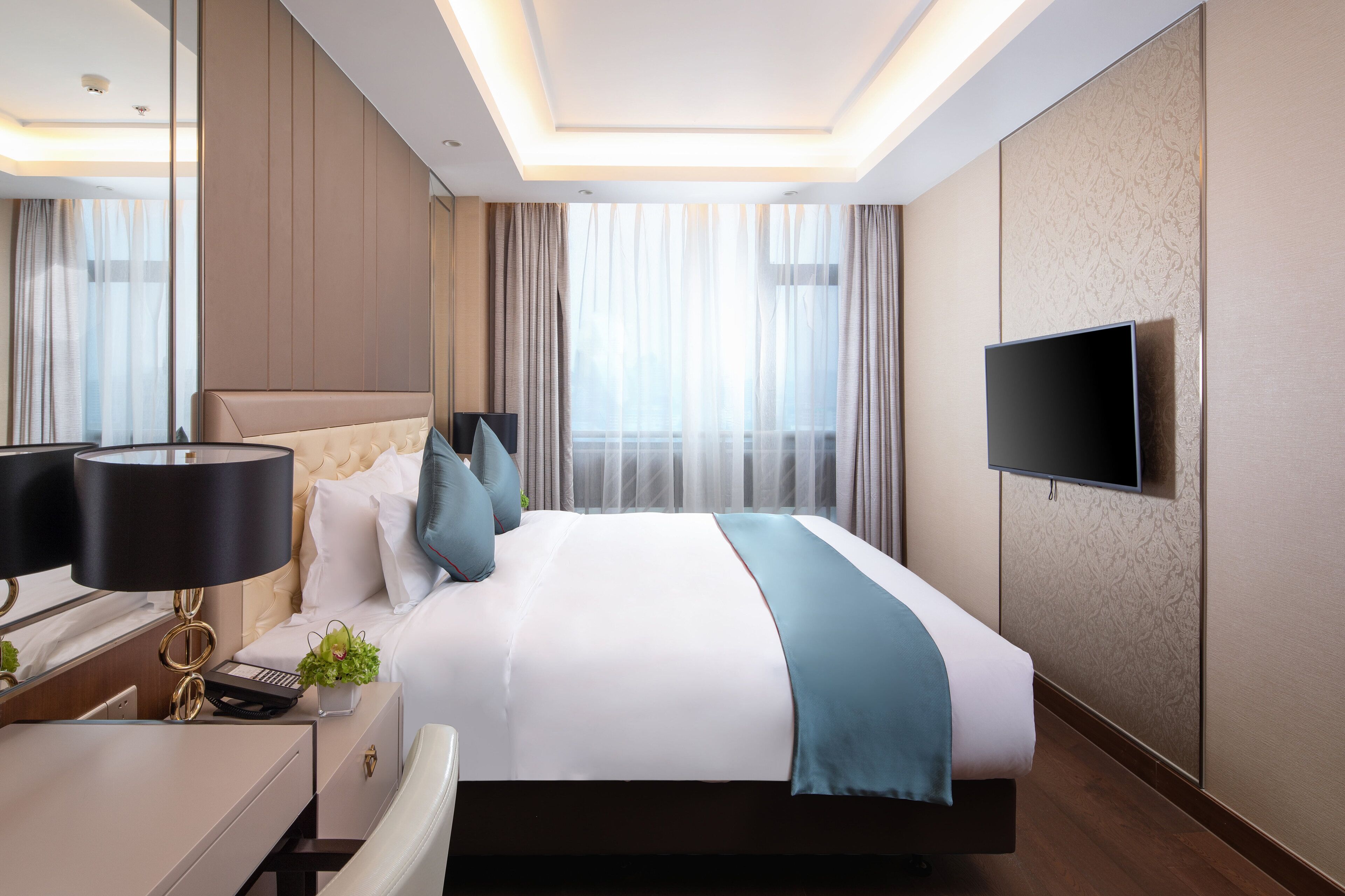 A modern hotel room with a large bed, a flat-screen TV mounted on the wall, a bedside table with a lamp, and a view of the city through large windows.
