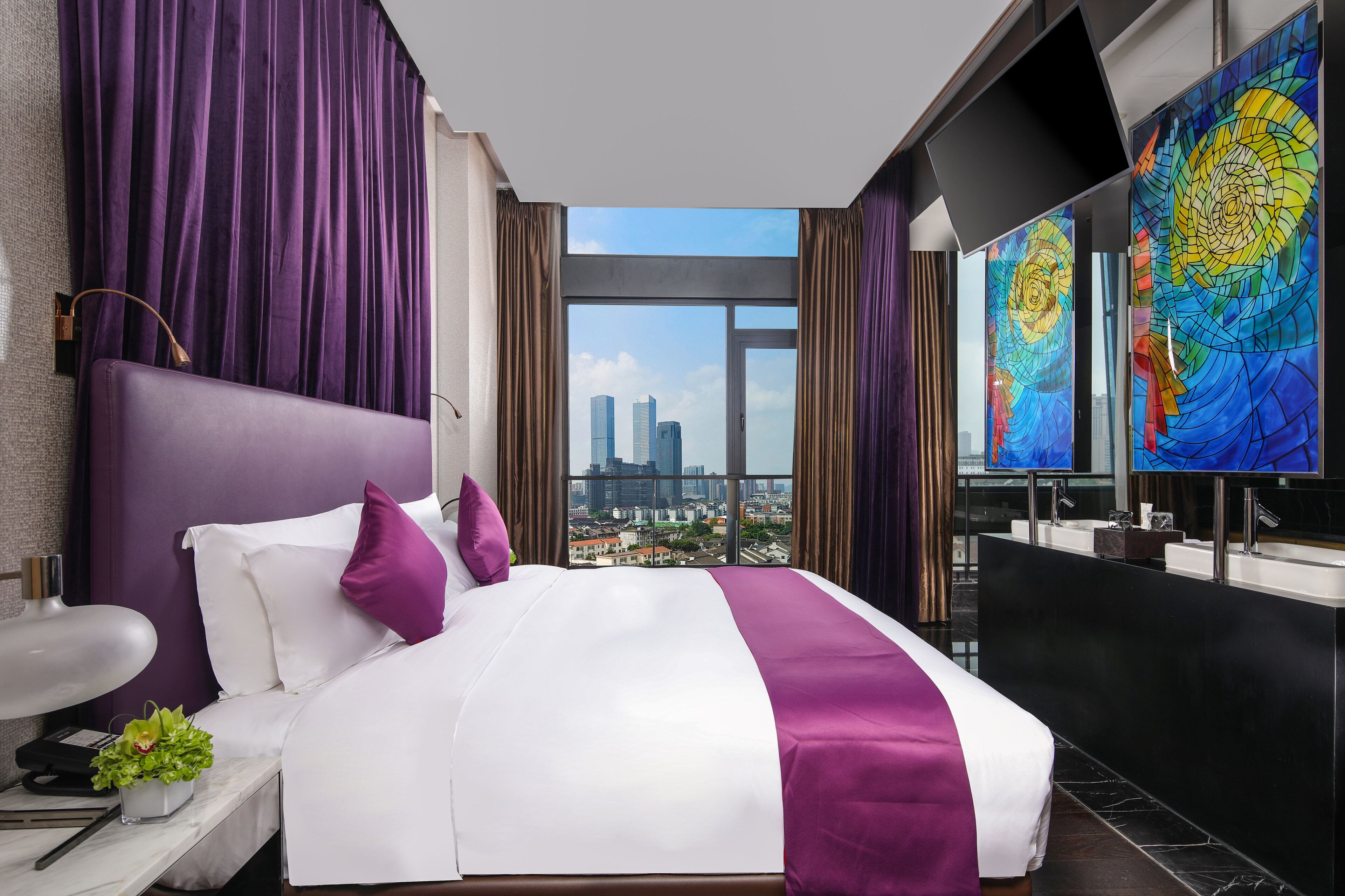 Una habitación de hotel moderna con una cama grande, cortinas moradas, vistas a la ciudad y un baño con un espejo amplio y obras de arte coloridas.