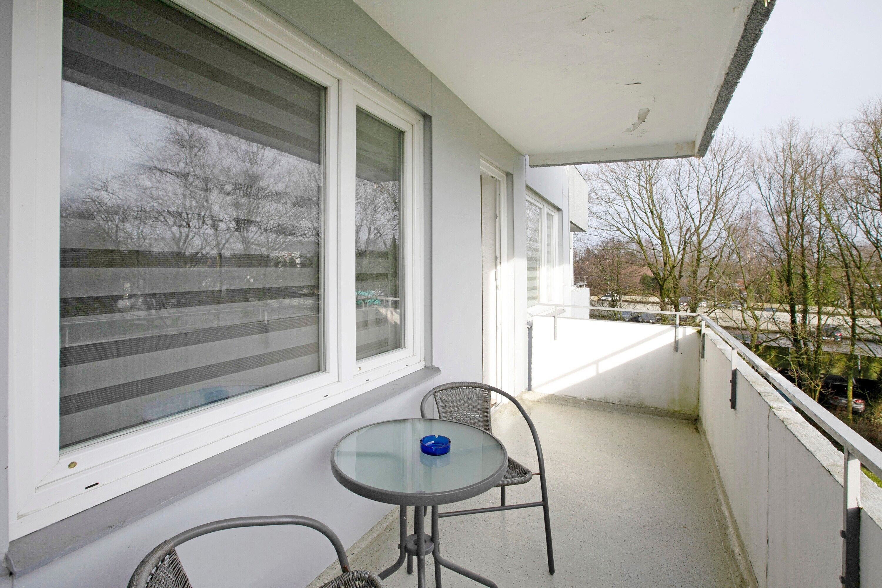 Un balcon avec une table et des chaises, de grandes fenêtres et une vue sur les arbres et les bâtiments.