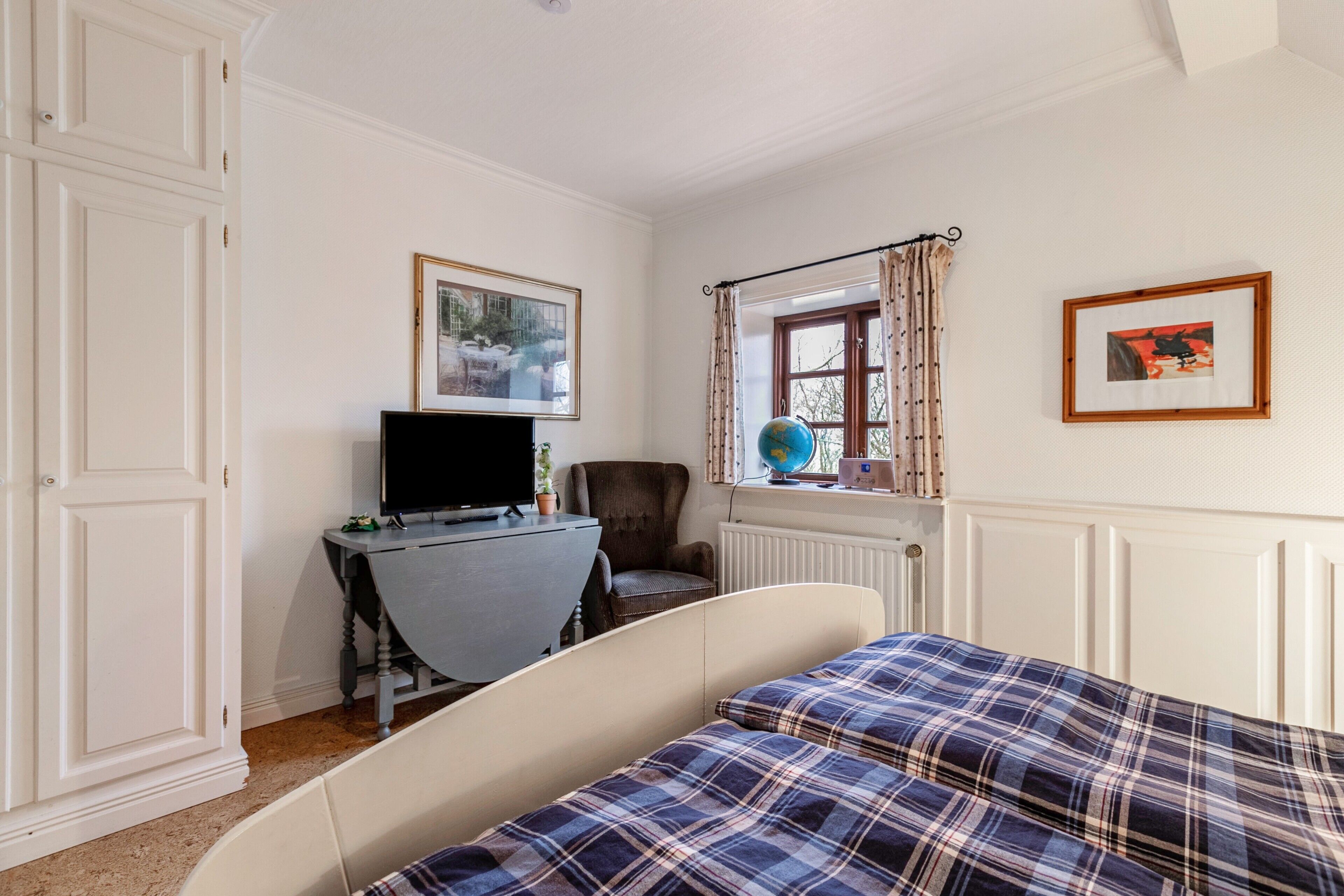 A bedroom with a bed, a desk with a computer, a chair, a globe, and framed pictures on the wall.