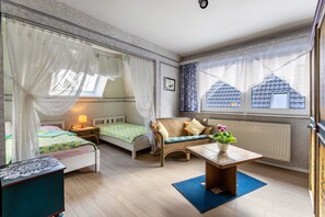 A room with two beds, a wicker chair, a table with a plant, and a view of buildings through large windows. - Apartment 'Erholung Pur' with Balcony and Wi-Fi (Hennstedt)