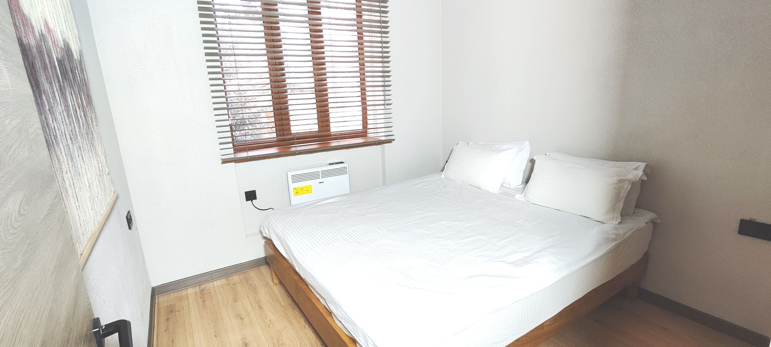 A small, neatly made bed in a room with a wooden floor and white walls. There is a window with blinds, a wall-mounted heater, and a door visible.