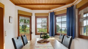 Ein Essbereich mit einem Holztisch, vier Stühlen, einer Tischläufer mit Blumenmuster und Blick ins Freie durch große, mit Vorhängen versehene Fenster.