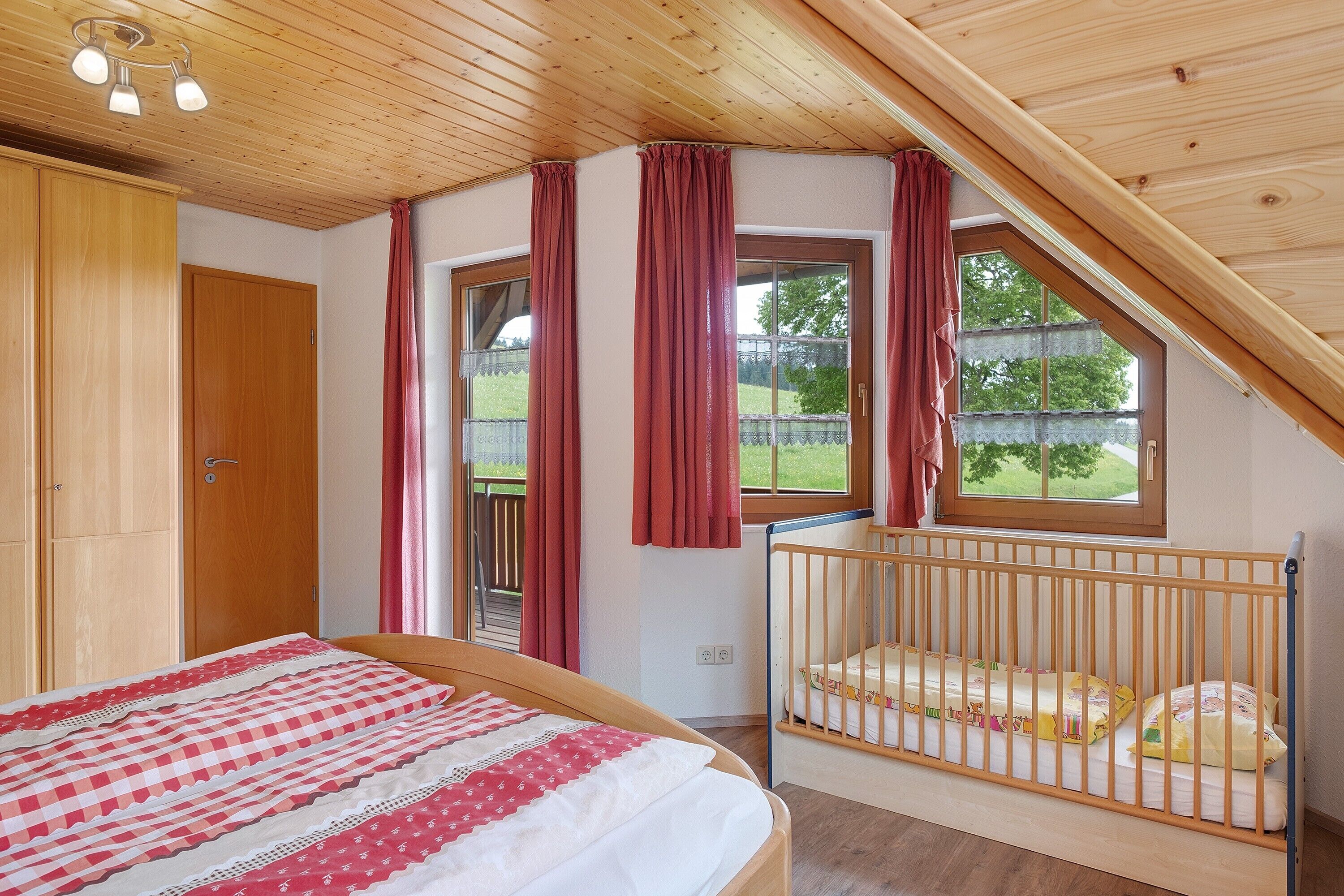 Ein Schlafzimmer mit Holzboden, einem Babybett, einem Bett mit kariertem Überwurf und einem Fenster mit Vorhängen.