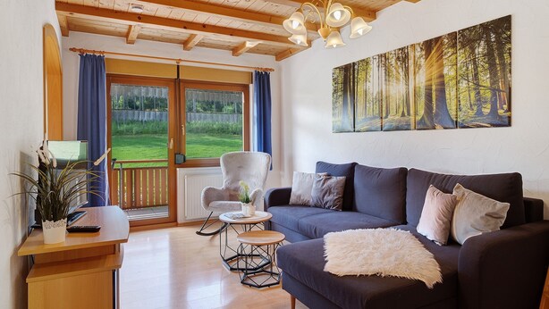 Ein Wohnzimmer mit einem dunklen Sofa, einem weißen Sessel, einem kleinen Holztisch und einem großen Waldgemälde an der Wand.