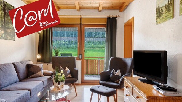 A living room with a sofa, two armchairs, a coffee table with flowers, a TV, and a sliding glass door to a balcony. - Holiday Apartment 'West' with Shared Terrace, Shared Garden and Balcony (Eisenbach (Hochschwarzwald))