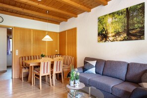 Une pièce avec un plafond en bois, une table à manger avec des chaises, un canapé gris et un tableau représentant une forêt.