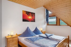 A bedroom with a sloped ceiling, a bed with blue bedding, two bedside lamps, and a painting of a sunset. - Holiday Apartment 'West' with Shared Terrace, Shared Garden and Balcony (Eisenbach (Hochschwarzwald))