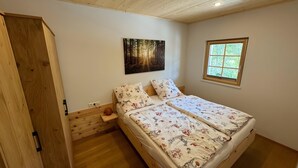 Een slaapkamer met een houten plafond, een bed met bloemenbeddengoed, wanddecoratie met een bosmotief en een raam met uitzicht op groen.