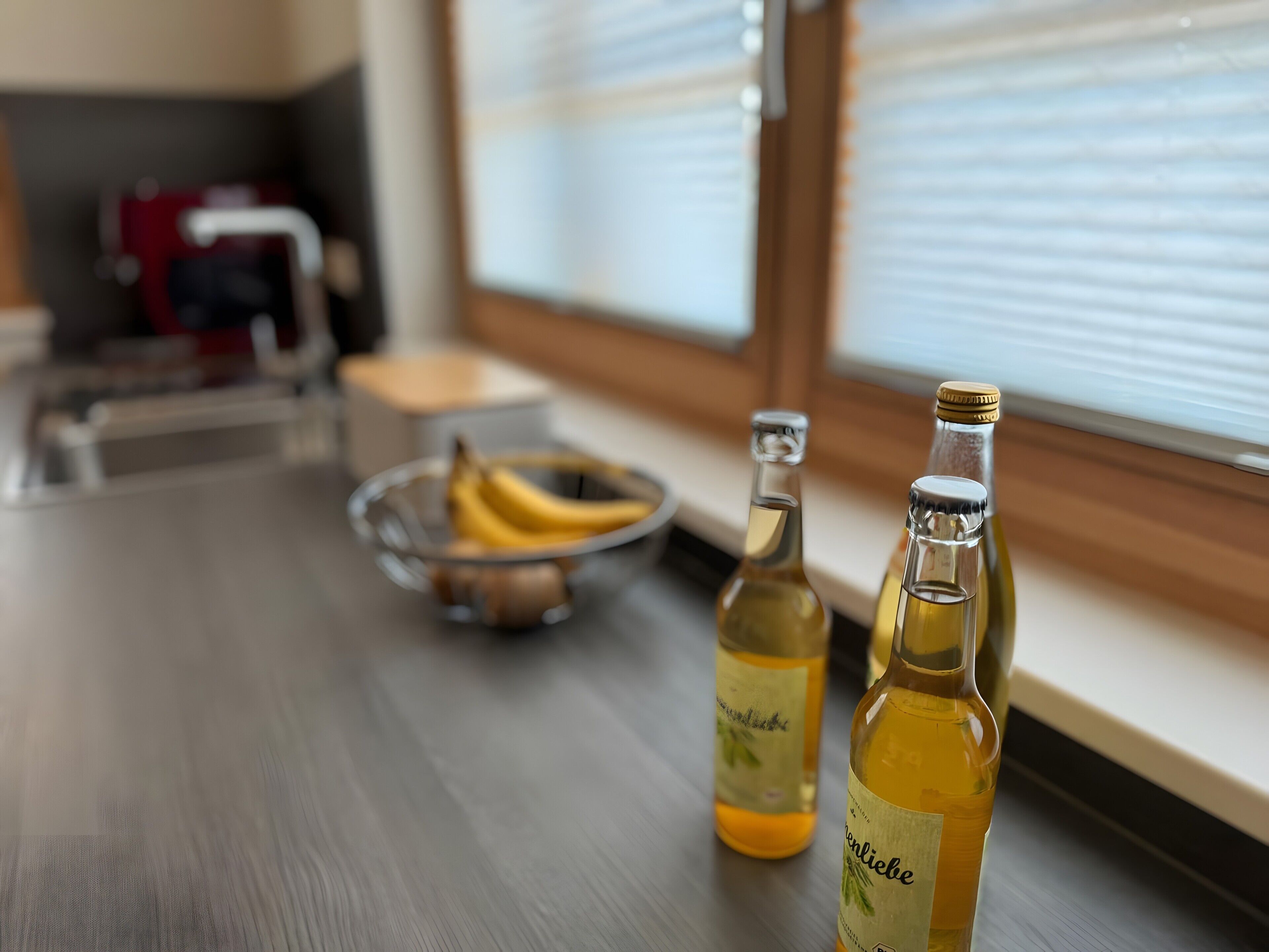 Een aanrecht met twee flessen wijn, een schaal bananen en een raam met zonwering.