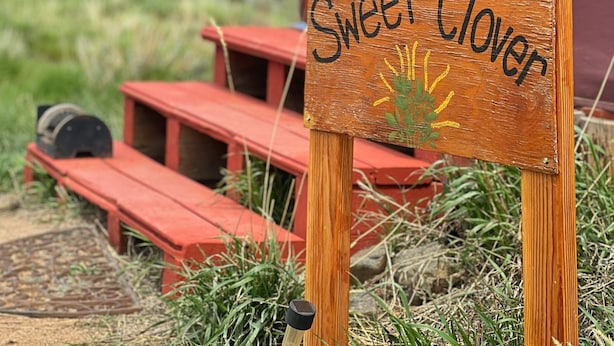 Un letrero de madera con la inscripción "Sweet Clover", ubicado frente a un banco rojo y una puerta de madera.