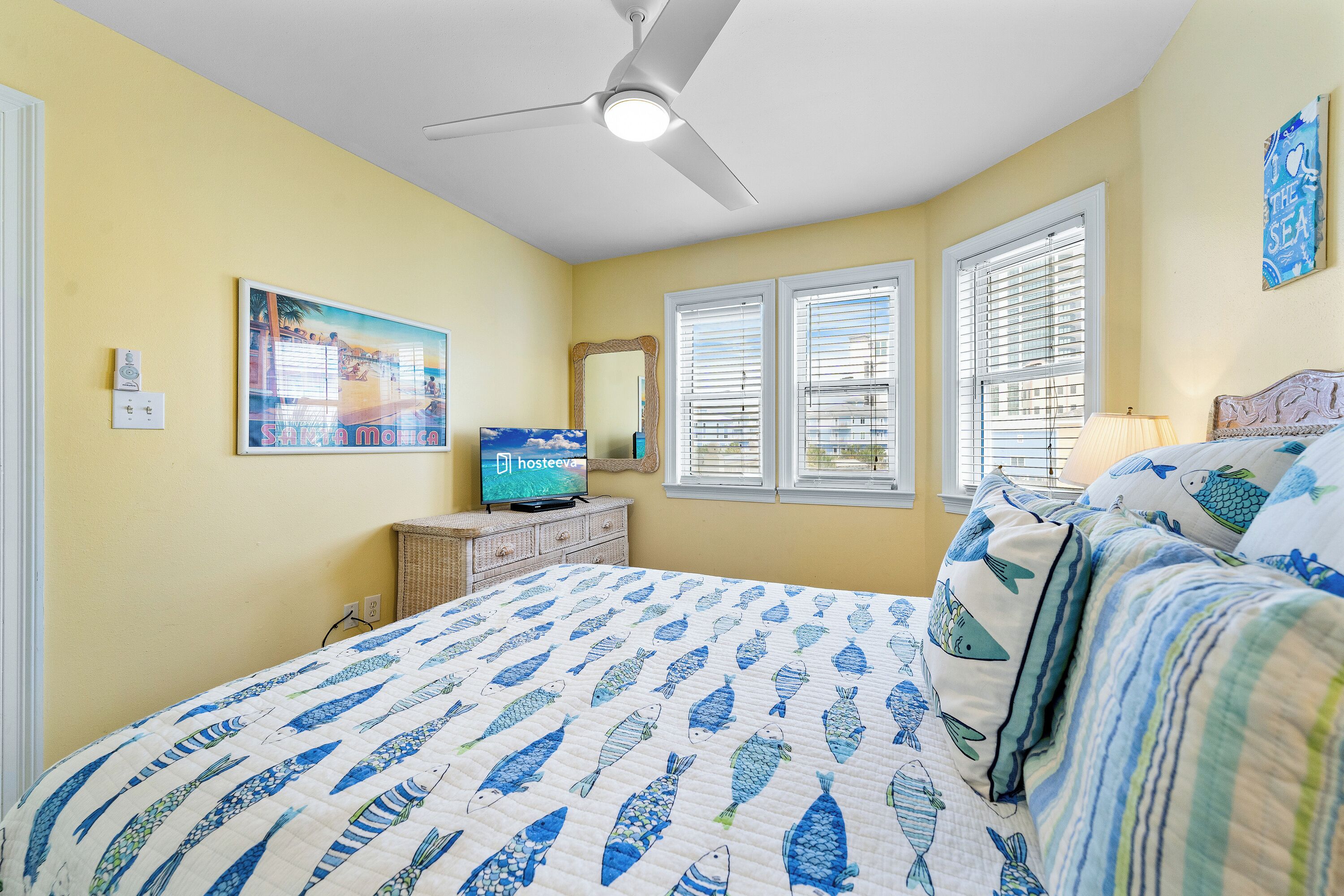 Une chambre à coucher comprenant un lit, un ventilateur de plafond, un téléviseur et une fenêtre avec des stores.