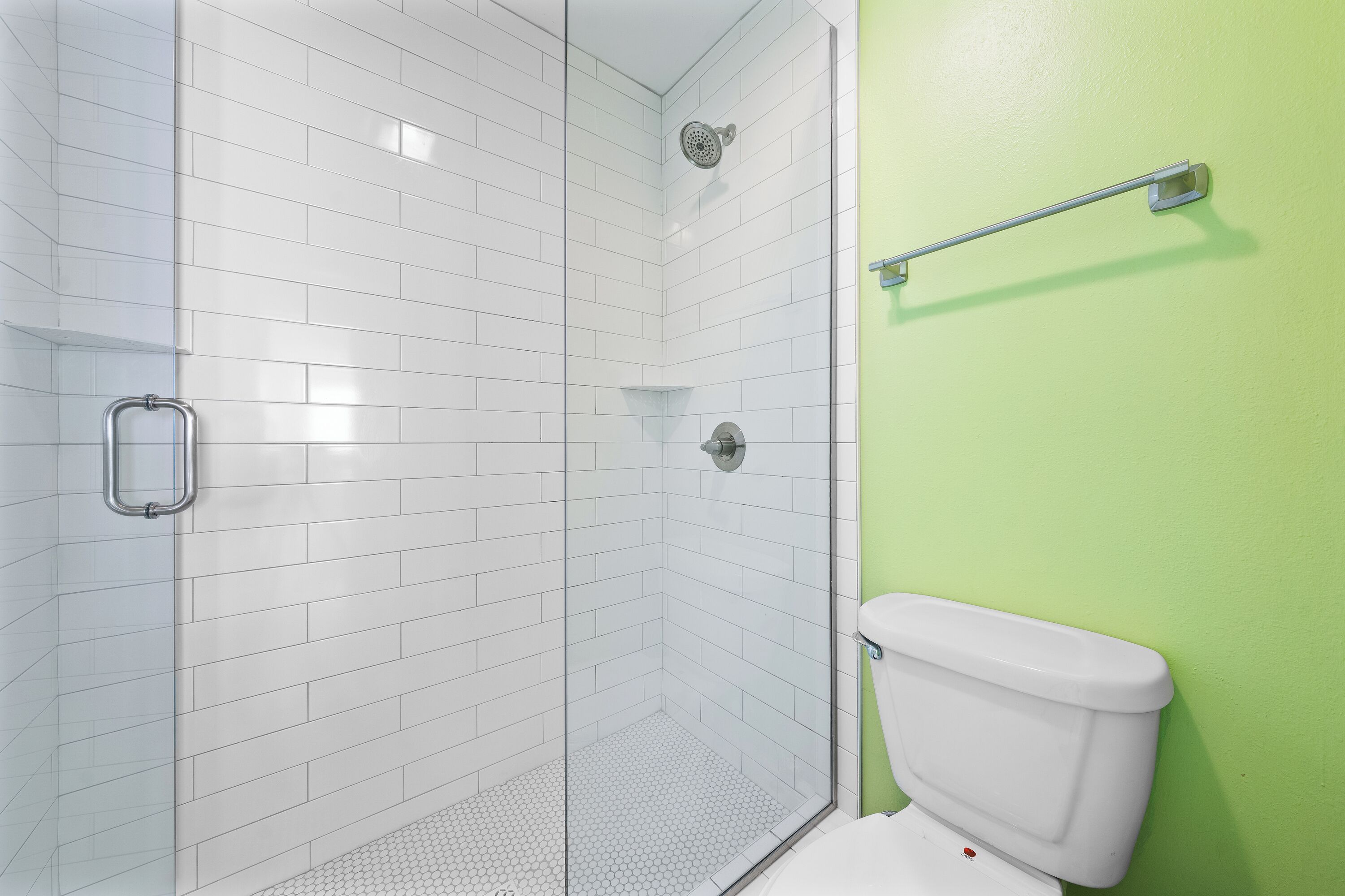 Ducha con azulejos blancos y puerta de vidrio, inodoro con pared verde y barra de apoyo metálica.