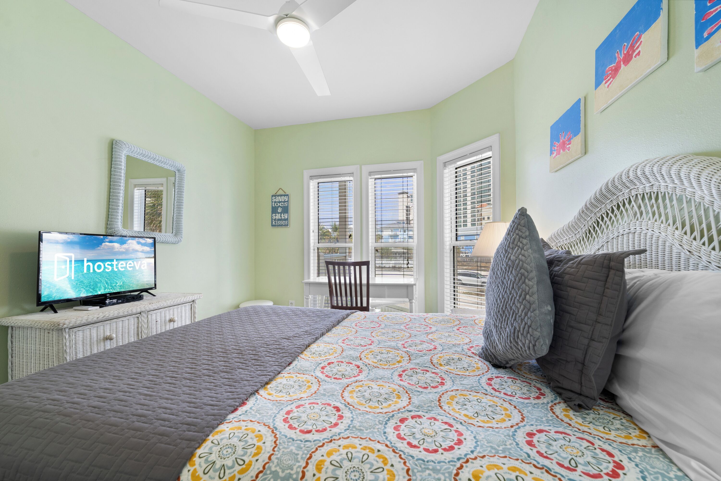 Un dormitorio con cama, televisor sobre un aparador blanco, ventana con persianas y ventilador de techo.