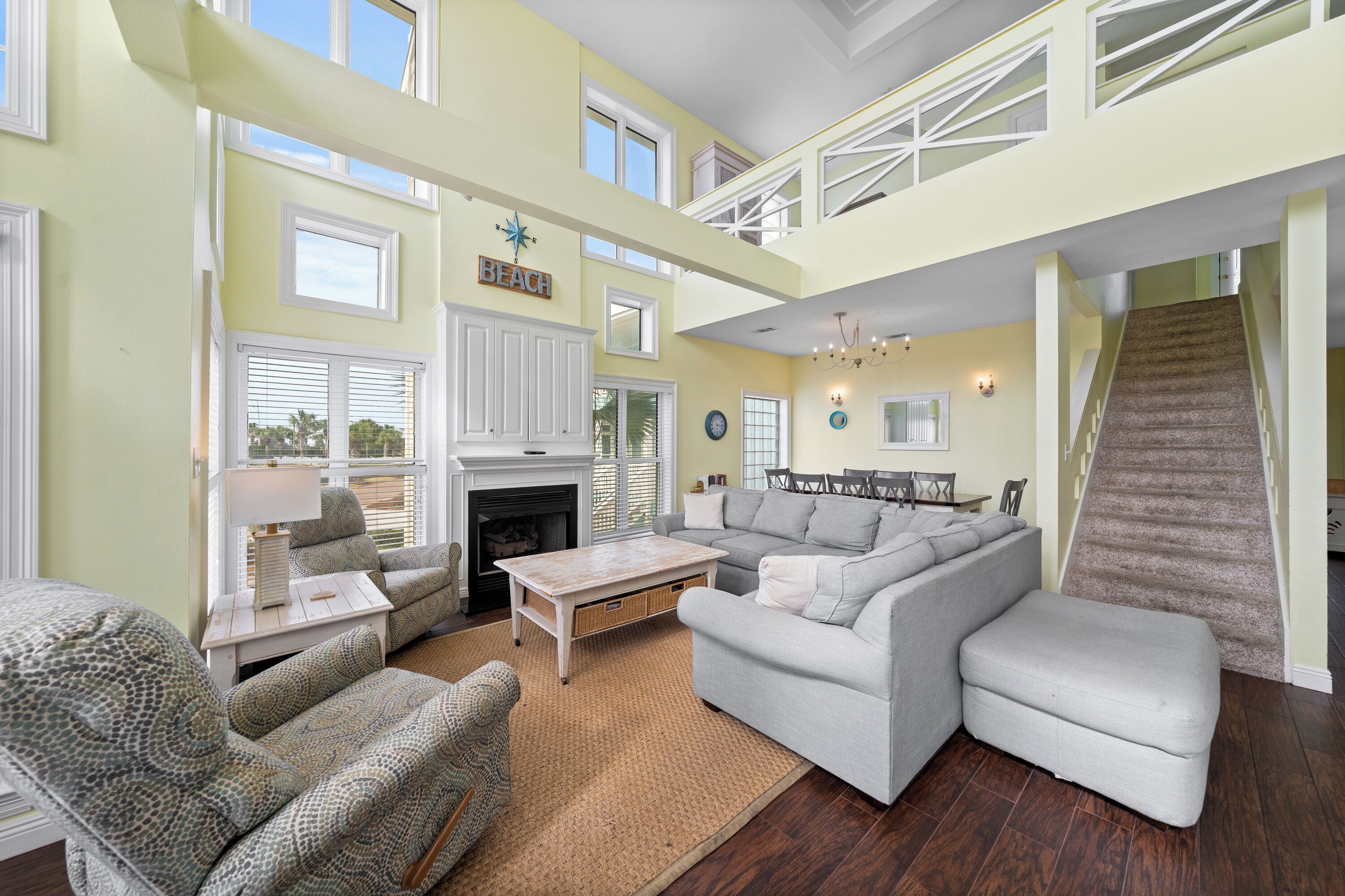 A spacious living room with a fireplace, a staircase, and a 'BEACH' sign on the wall.