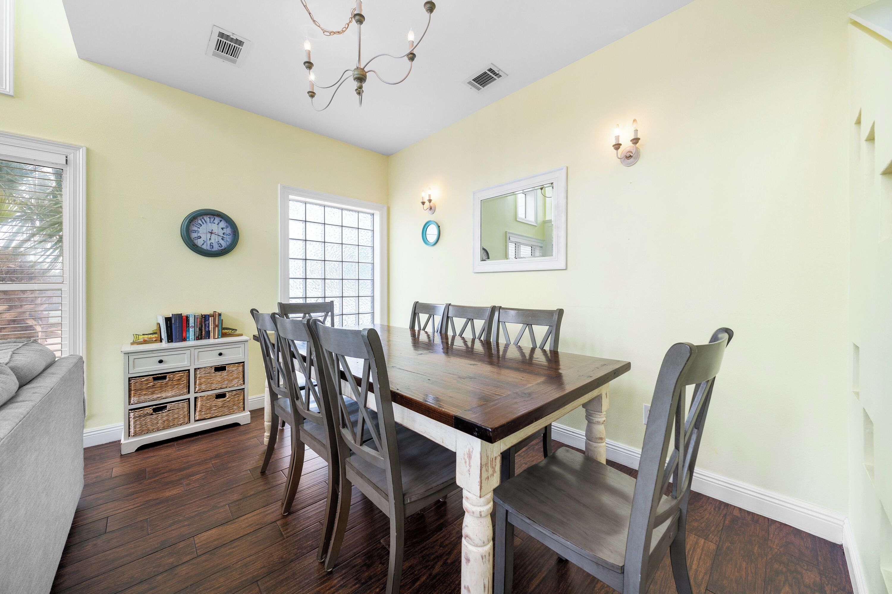 A dining area with a wooden table and chairs, a white sideboard with baskets, a wall clock, and a window with blinds.