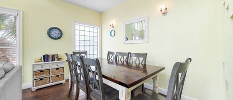 A dining area with a wooden table and chairs, a white sideboard with baskets, a wall clock, and a window with blinds.