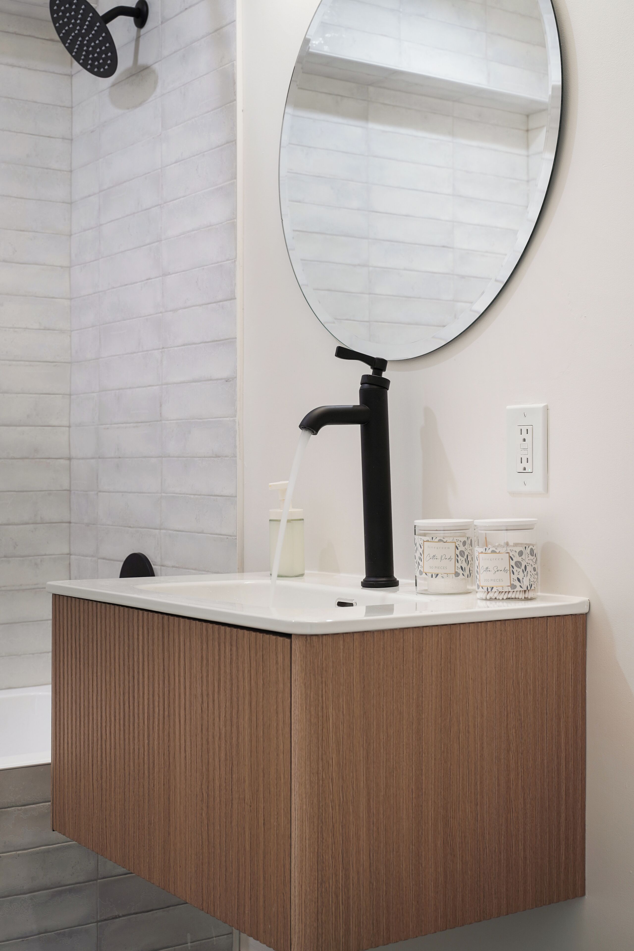 A bathroom with a wooden vanity, a black faucet, a round mirror, and a showerhead.