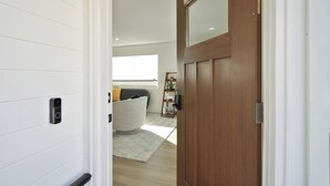 Un corridoio con una porta in legno, una parete bianca con telecamera di sicurezza, un divano grigio e una finestra con persiane.