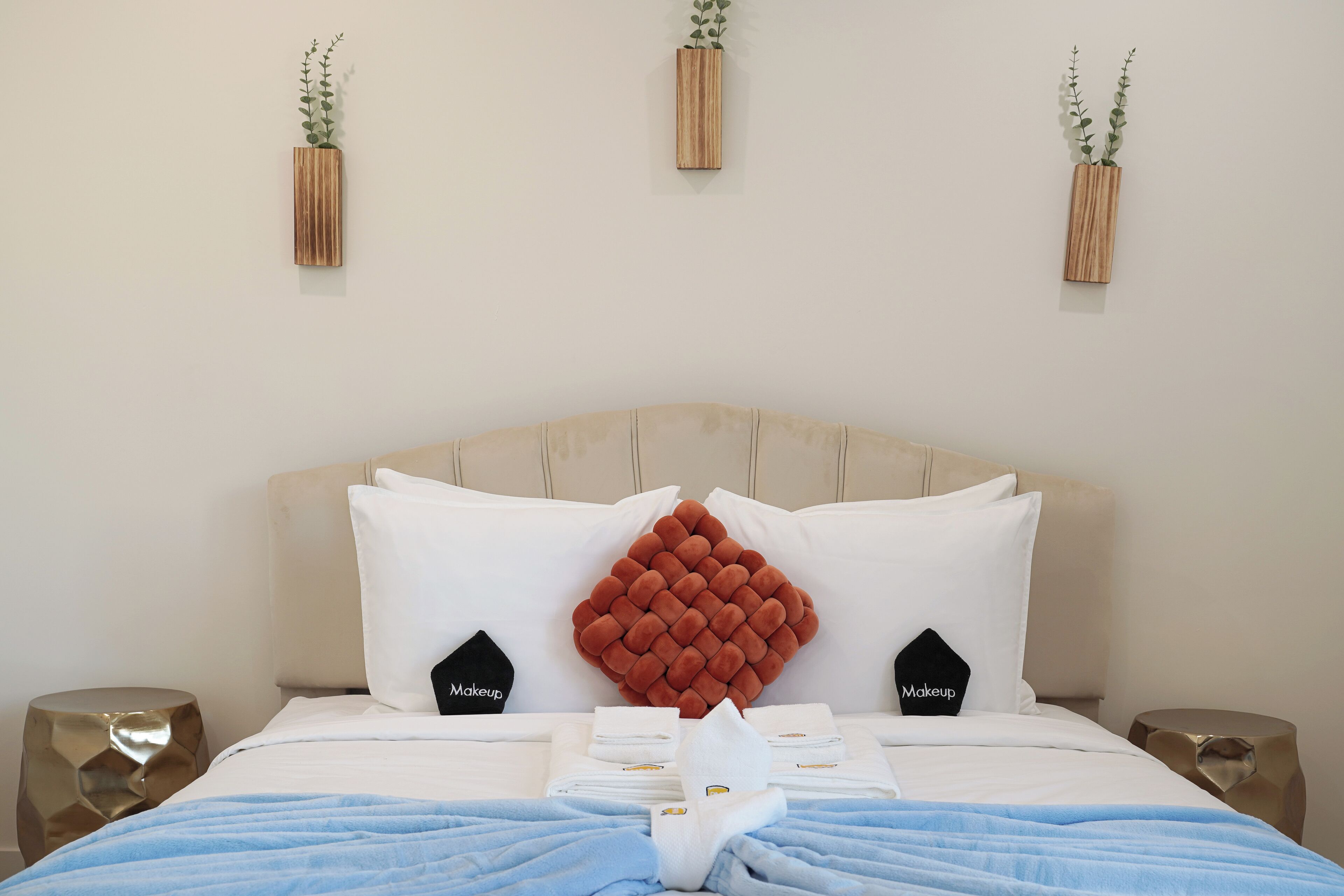 Una cama bien hecha con un llamativo cabecero tapizado rojo, almohadas blancas y una manta azul. En la pared sobre la cama, hay dos pequeños jarrones de madera con plantas verdes.