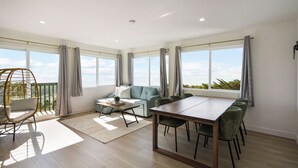 A spacious living room with a sofa, a dining table, and chairs, large windows with curtains, and a balcony with a wicker chair. - Oceanview Sunset View 3BR Luxe Home Yard (Pacifica)
