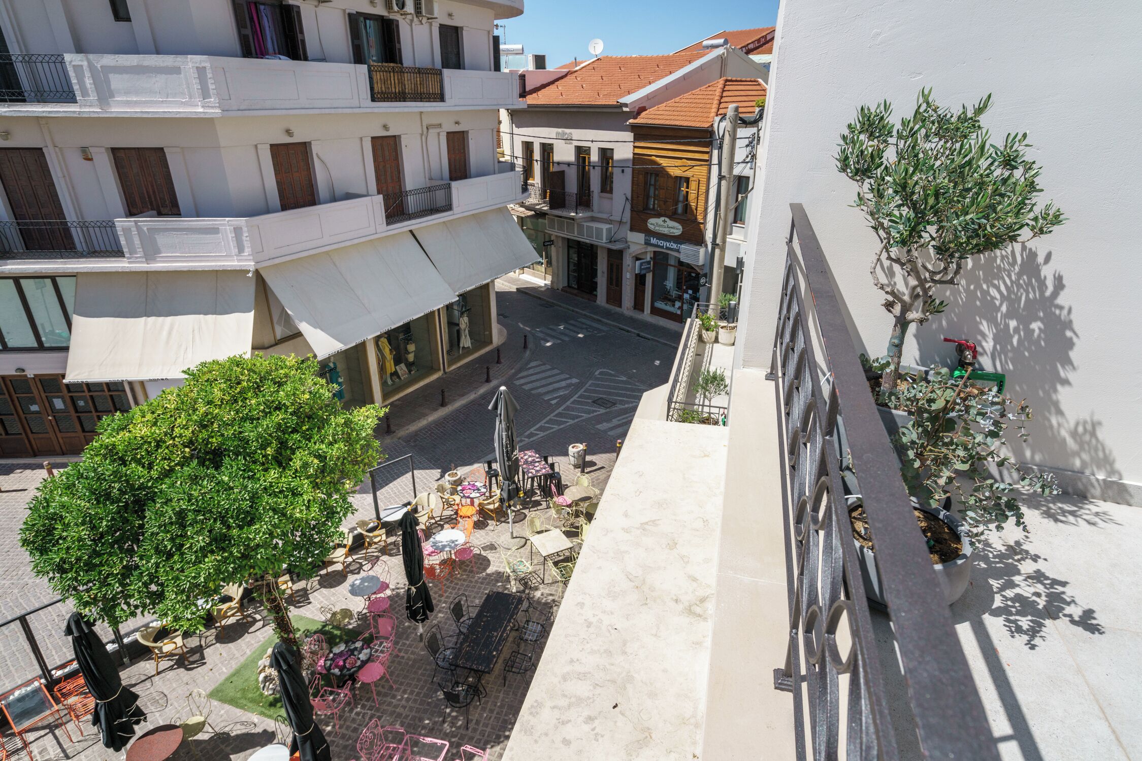 An outdoor cafe area with tables and chairs, a tree, and a building with balconies and shops in the background.