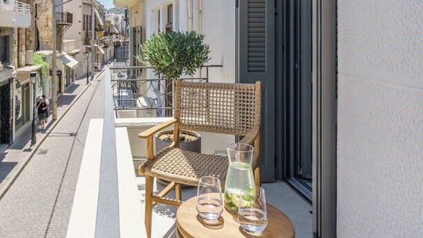 A narrow street with a balcony featuring a wooden table, two wicker chairs, and a potted plant. - The Old Distillery Premium Suites (Rethymno)