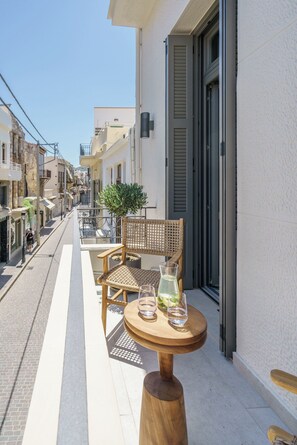 A narrow street with a balcony featuring a wooden table, two wicker chairs, and a potted plant. - The Old Distillery Premium Suites (Rethymno)