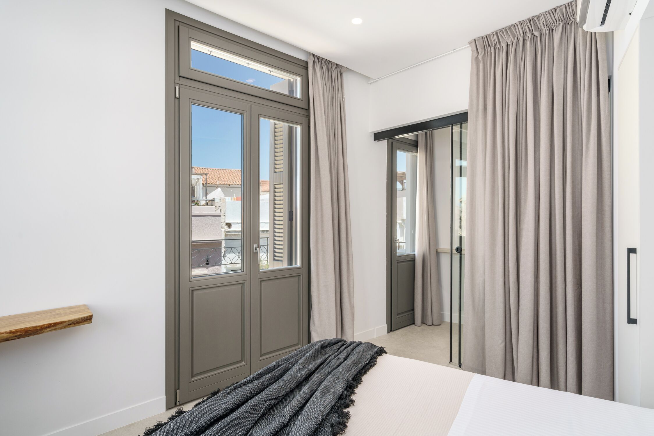 A bedroom with a large window, a bed with a grey blanket, a wooden shelf, and a wardrobe.