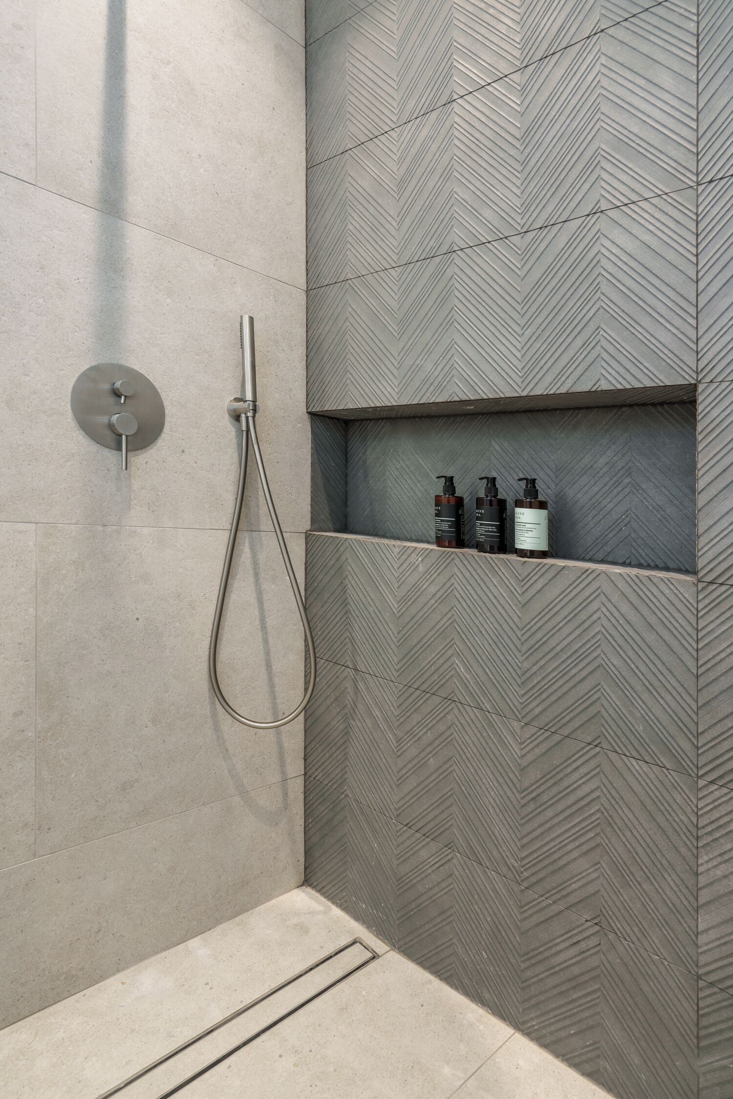A shower area with a herringbone-patterned wall, a handheld showerhead, and a shelf with toiletries.