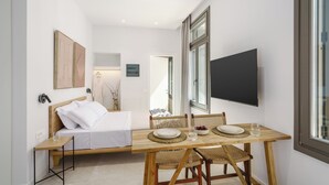 A compact room with a bed, a dining table with chairs, a TV mounted on the wall, and a window. - The Old Distillery Premium Suites (Rethymno)