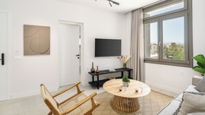 A modern living room with a round wooden table, two wicker chairs, a black entertainment unit, and a large window with a city view. - The Old Distillery Premium Suites (Rethymno)