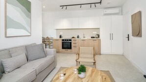 A modern living room with a sofa, coffee table, and a chair. A kitchen with white cabinets and a dining area is visible in the background. - The Old Distillery Premium Suites (Rethymno)