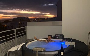 Une personne se détend dans un bain à remous, avec une vue sur le coucher de soleil, une bouteille de vin et une télécommande à portée de main.