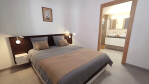 A bedroom with a bed, bedside tables, a nightstand, a framed picture on the wall, and a bathroom visible through an open door. - T4 duplex and rooftop in the heart of Saint-Pierre, this new luxury residence be (Saint-Pierre)