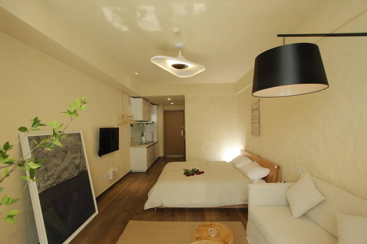 Une chambre à coucher moderne avec un lit, un canapé blanc, une lampe noire suspendue et un petit coin cuisine à l’arrière-plan.