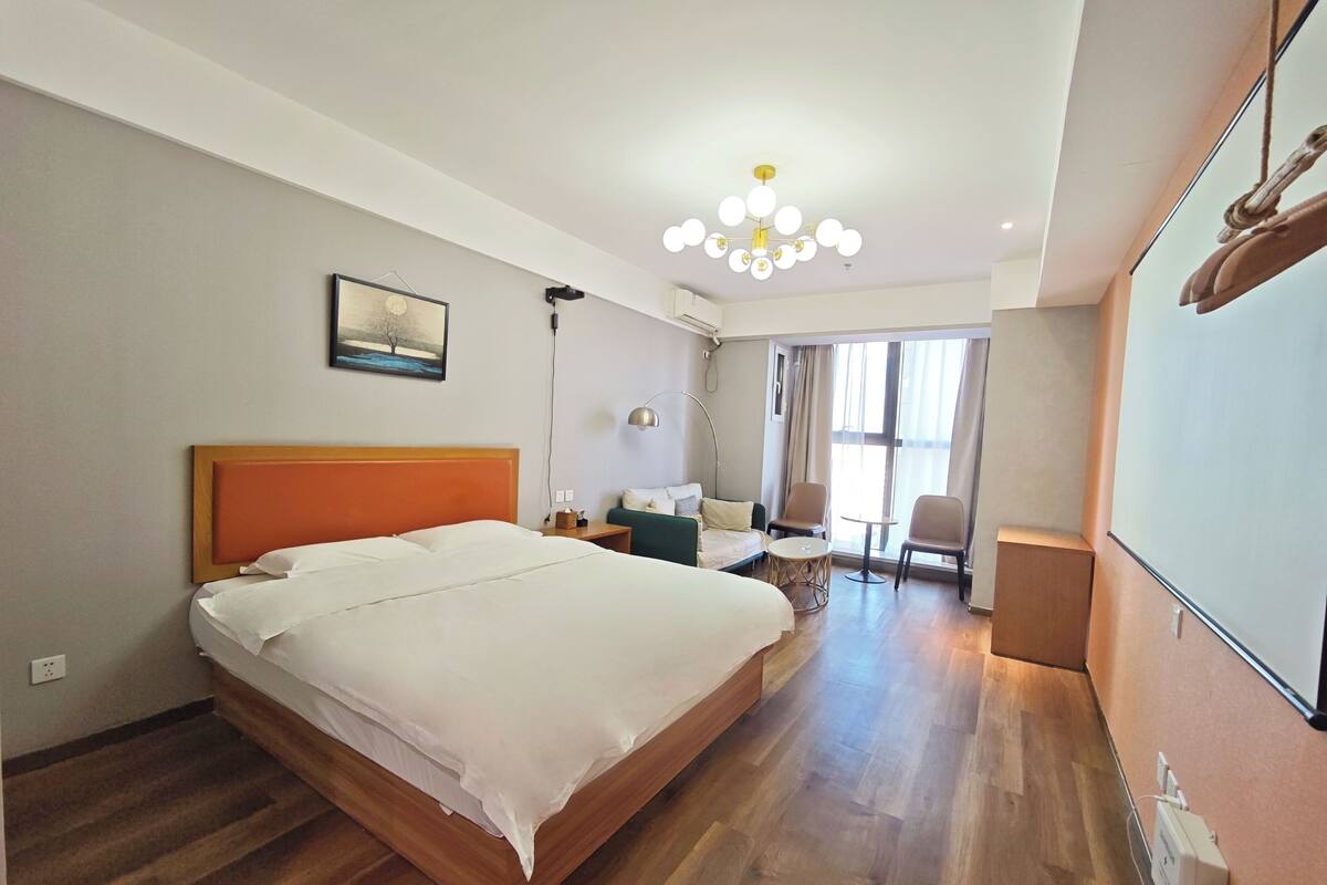 Une chambre d’hôtel avec un grand lit, un bureau, deux chaises, une petite table et une grande fenêtre avec des rideaux.