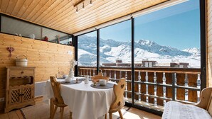 A wooden interior with a table set for dining, a wooden cabinet, and a view of snow-covered mountains through large windows.
